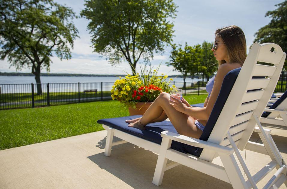 Single woman sitting on a lounge chair next to a pool. She's wearing a violet bathing suit and drinking a glass of water. She's looking out towards the lake.