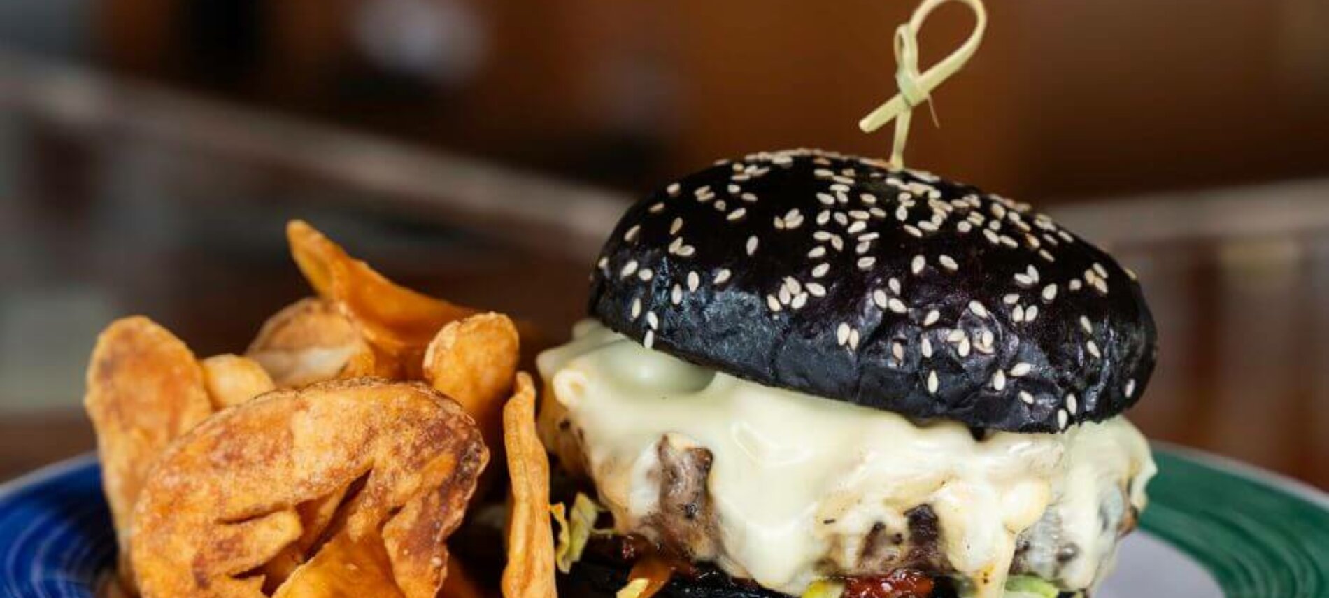 Delicious looking burger, with a black bun, burger, and mac'n'cheese oozing over the top of the burger. The burger has a side of chips.