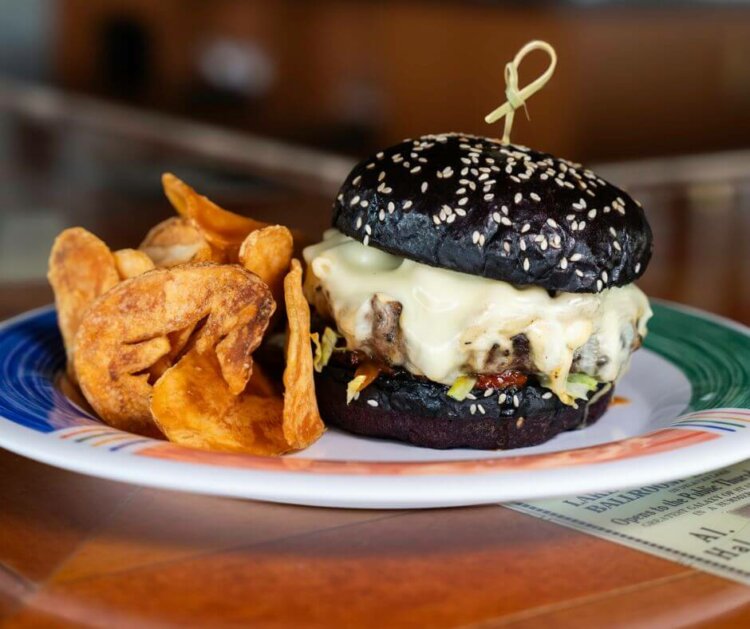 Delicious looking burger, with a black bun, burger, and mac'n'cheese oozing over the top of the burger. The burger has a side of chips.